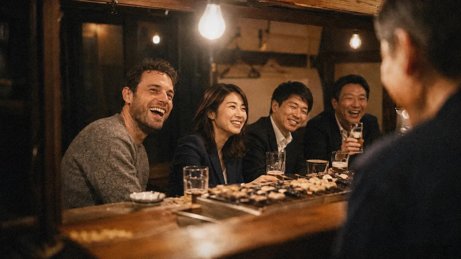 Guests laughing at the counter of a small Japanese izakaya, seen from behind the bar